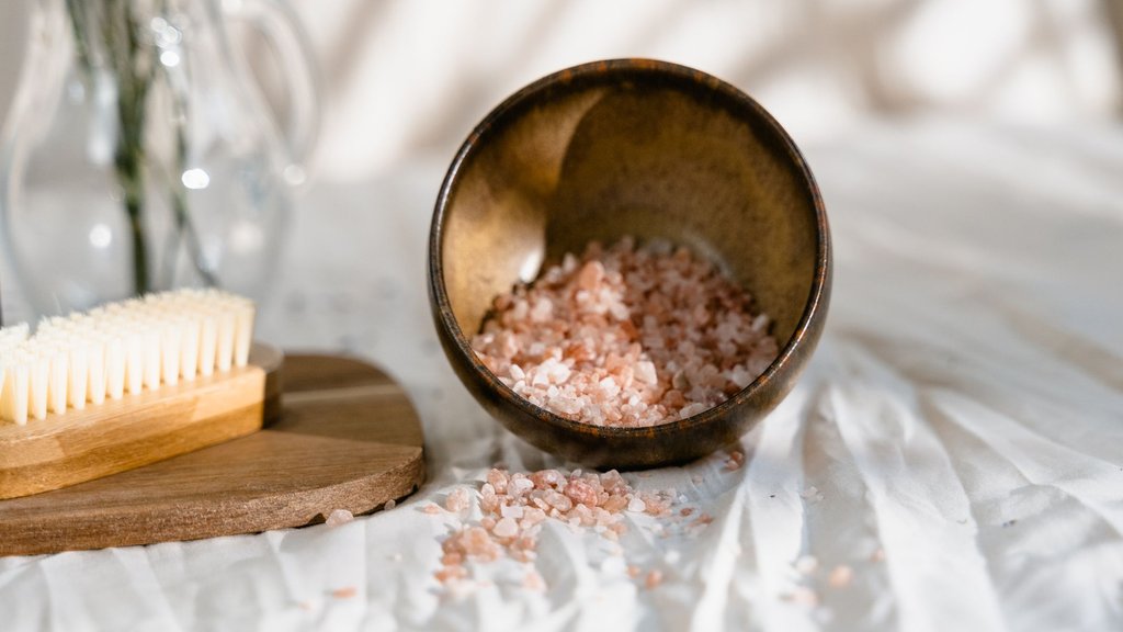 A rustic wooden bowl tipped on its side spilling pink Himalayan salt scrub crystals onto cream linen, beside a natural bristle body brush on a wooden board and a glass jug of dried sprigs, in soft warm sunlight.