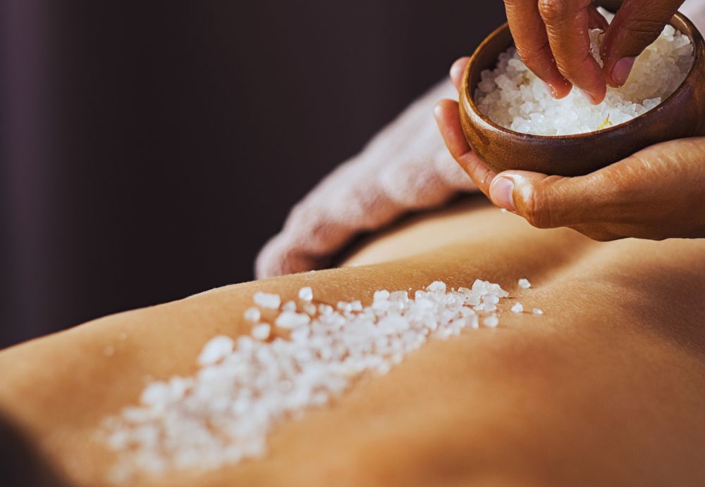 Coarse salt body scrub being sprinkled from a wooden bowl onto a client's back